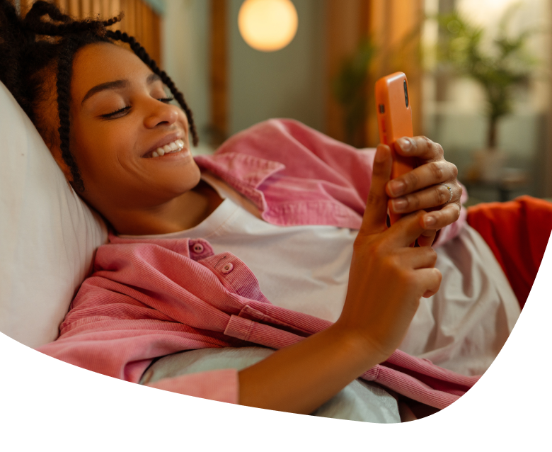 A young woman lies on a bed, smiling while looking at and using an orange smartphone. She is dressed casually in a pink shirt and white t-shirt, with soft, warm lighting in the background.