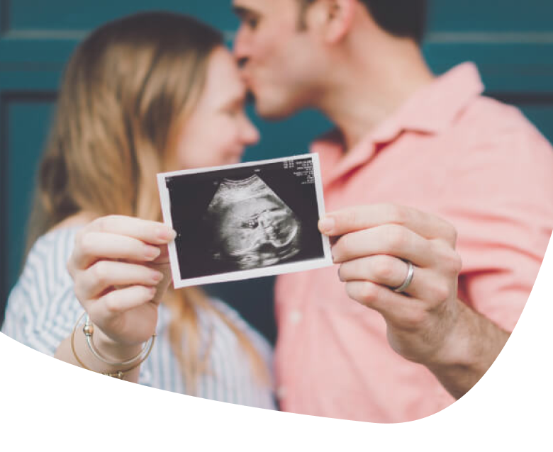 A couple stands close together, with the man kissing the womans forehead. They hold an ultrasound photo, suggesting they are expecting a baby. Their faces are out of focus, but the ultrasound is clearly visible.