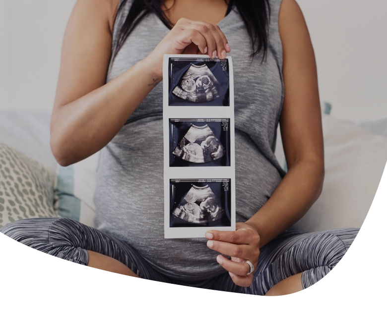 A pregnant woman in a gray outfit sits cross-legged on a bed, holding three ultrasound images in front of her belly, partially obscuring her face.
