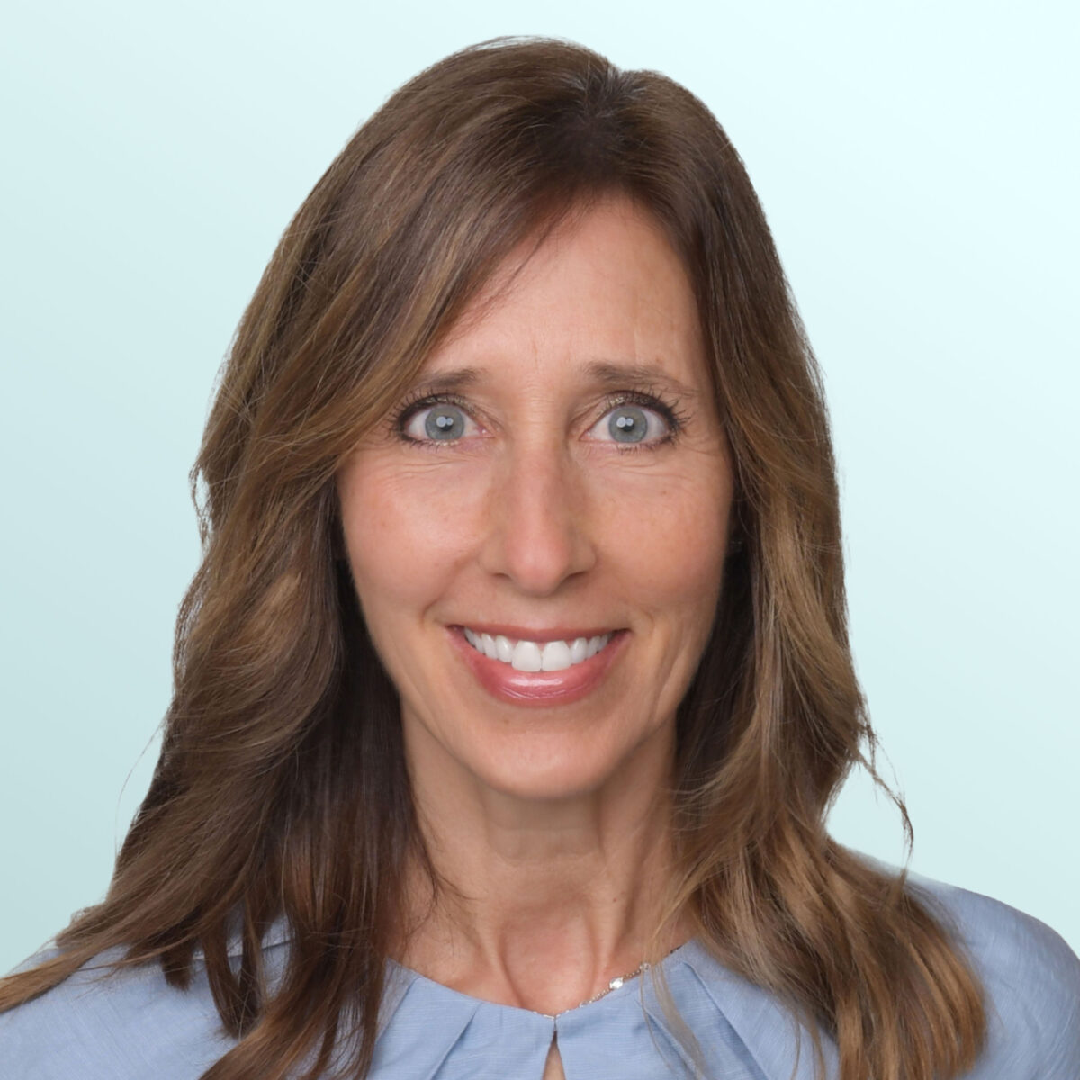 A woman with long brown hair and blue eyes smiles at the camera. She is wearing a light blue top and is posed against a pale blue background.