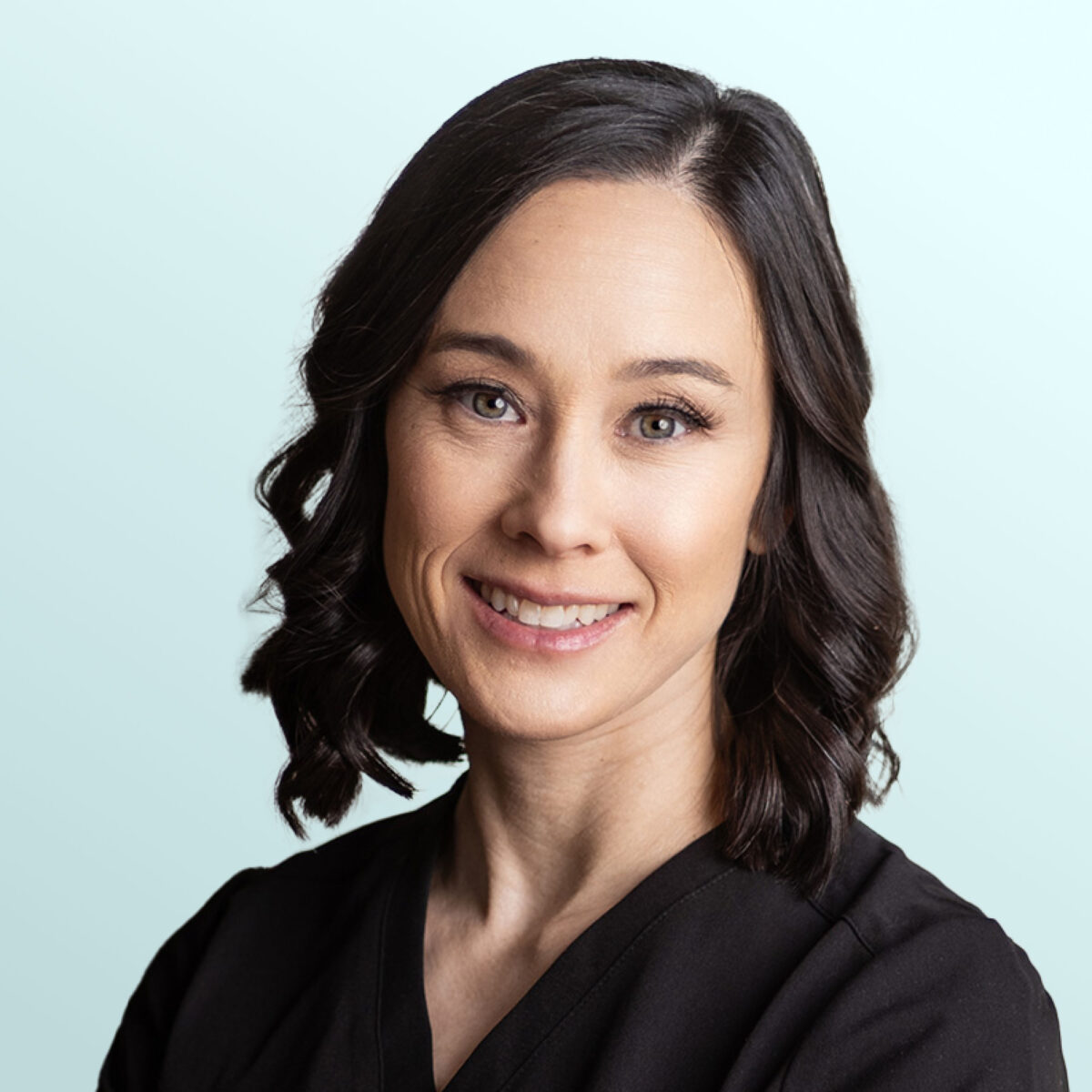 A woman with shoulder-length dark brown wavy hair, wearing a black top, smiles at the camera against a light blue background.