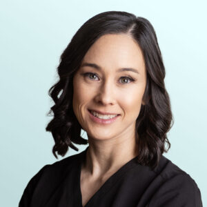 A woman with shoulder-length dark brown wavy hair, wearing a black top, smiles at the camera against a light blue background.