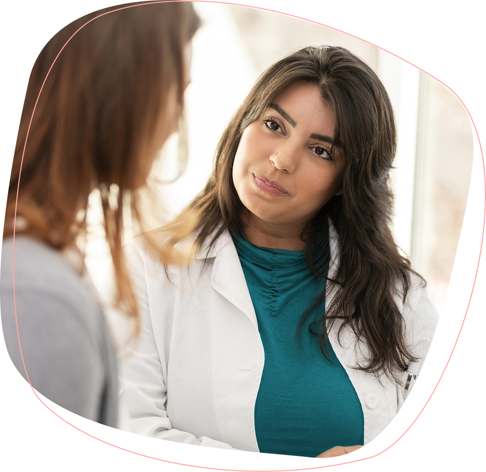 A female doctor with long dark hair, wearing a white coat and teal top, attentively listens to a patient during a consultation in a bright, softly lit room.