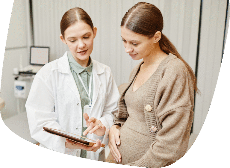 A doctor in a white coat shows a tablet to a pregnant woman wearing a brown sweater, who is smiling and holding her belly. They appear to be discussing something in a medical office.