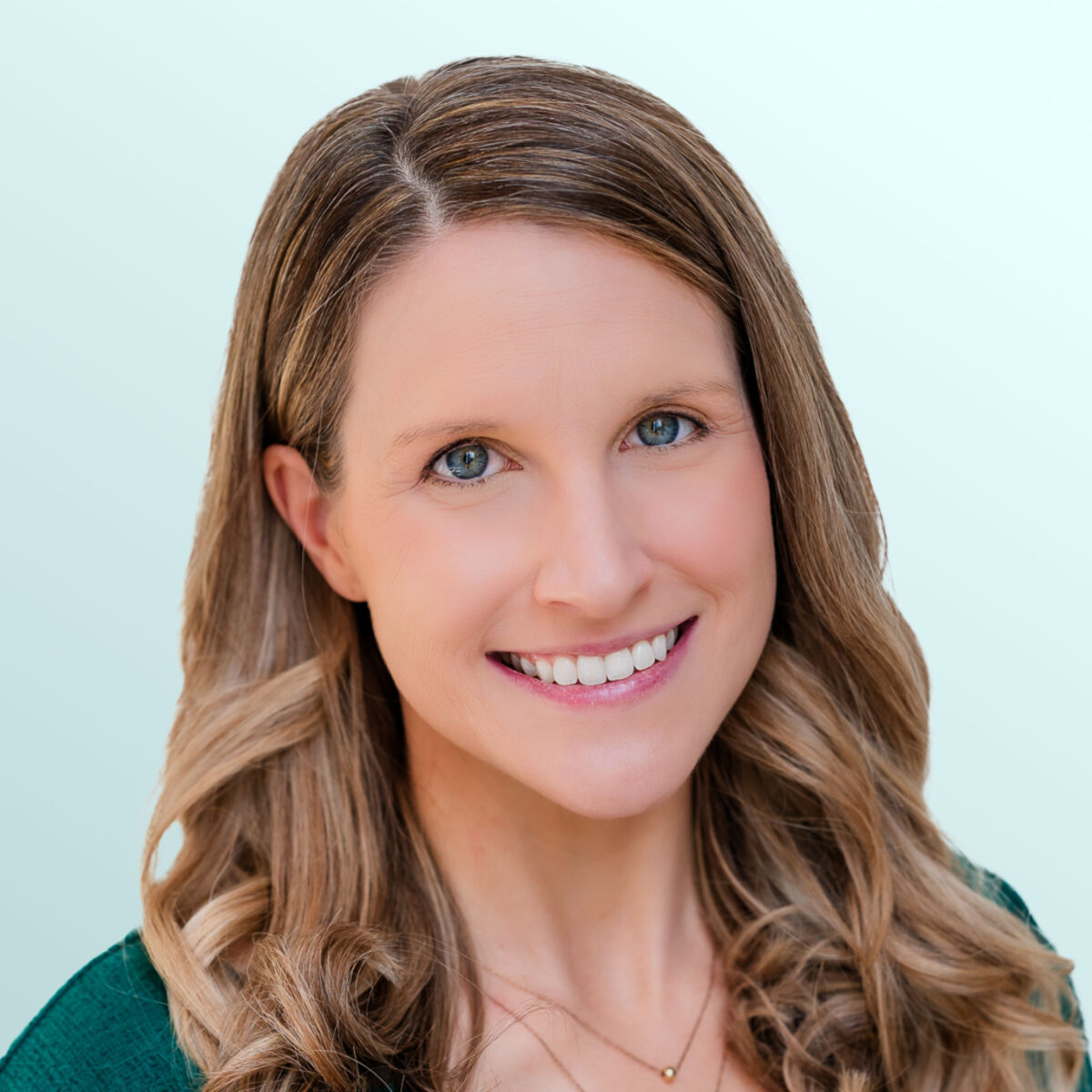 A woman with long, wavy light brown hair wearing a green top and two delicate necklaces smiles at the camera against a light blue background.