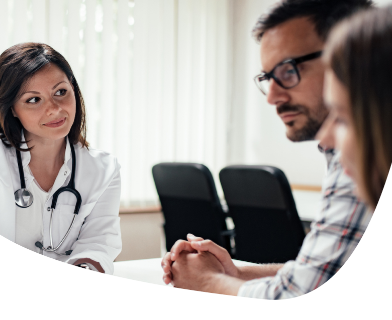 A female doctor with a stethoscope talks to a man and woman sitting across from her at a desk in a bright office, suggesting a medical consultation or discussion.