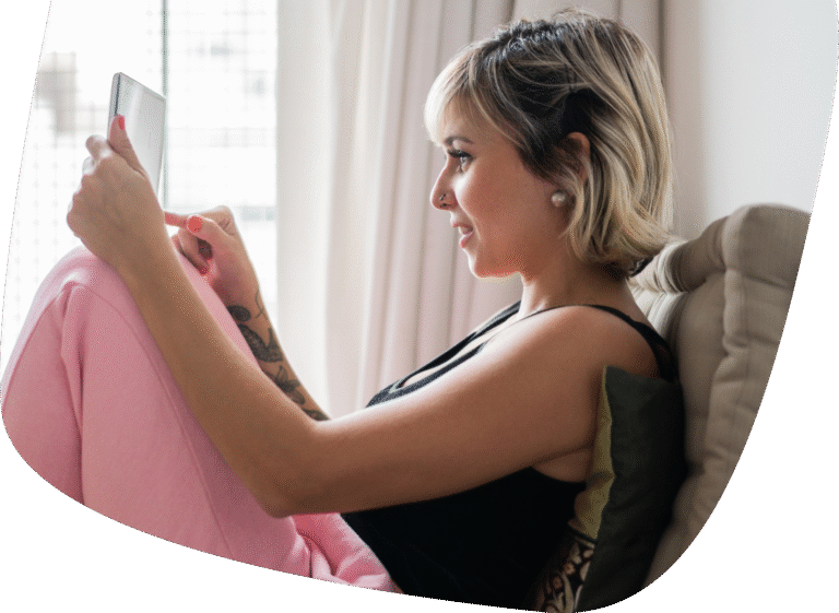 A woman with short blonde hair in a black top and pink pants sits on a couch, holding and looking at a tablet. She is relaxed, with her back against some cushions, near a window with sheer curtains.