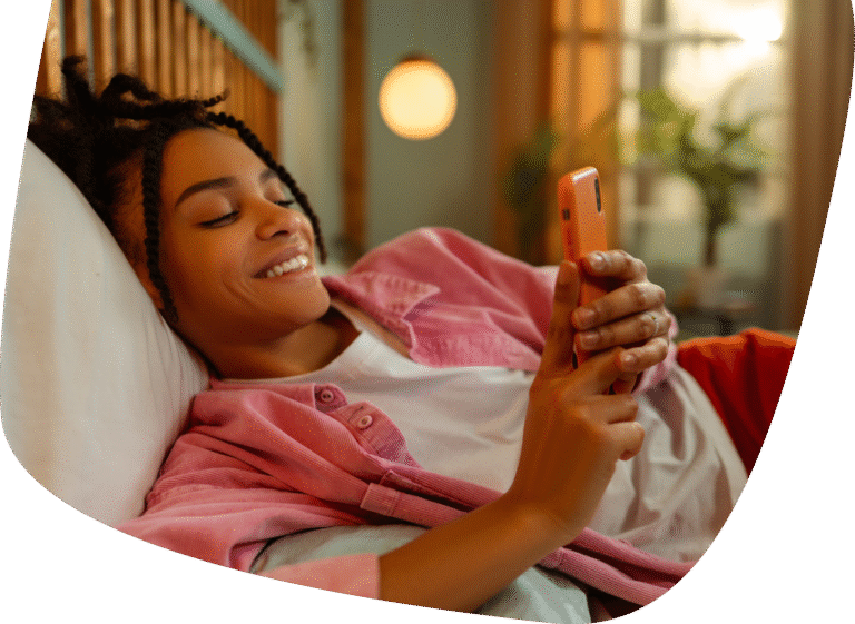 A person with braided hair is lying on a bed, smiling and looking at a smartphone. The background is cozy, with warm lighting and plants visible.