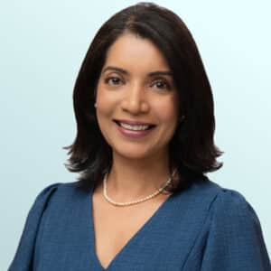 A woman with medium-length dark hair, wearing a blue dress and a pearl necklace, smiles at the camera against a light blue background.