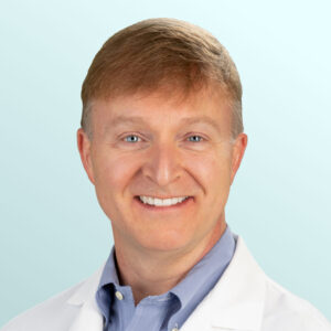 A smiling man with light brown hair wearing a white lab coat and a light blue collared shirt, posed in front of a light blue gradient background.