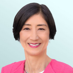 A woman with short black hair wearing a bright pink blazer and a silver necklace smiles at the camera against a light blue background.