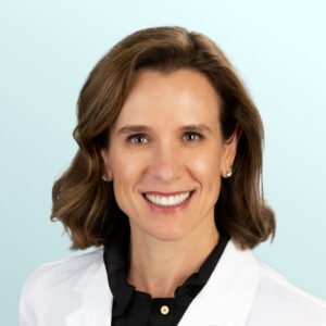 A woman with short brown hair smiles at the camera, wearing a white lab coat over a black shirt. She has stud earrings, and the background is a light blue gradient.