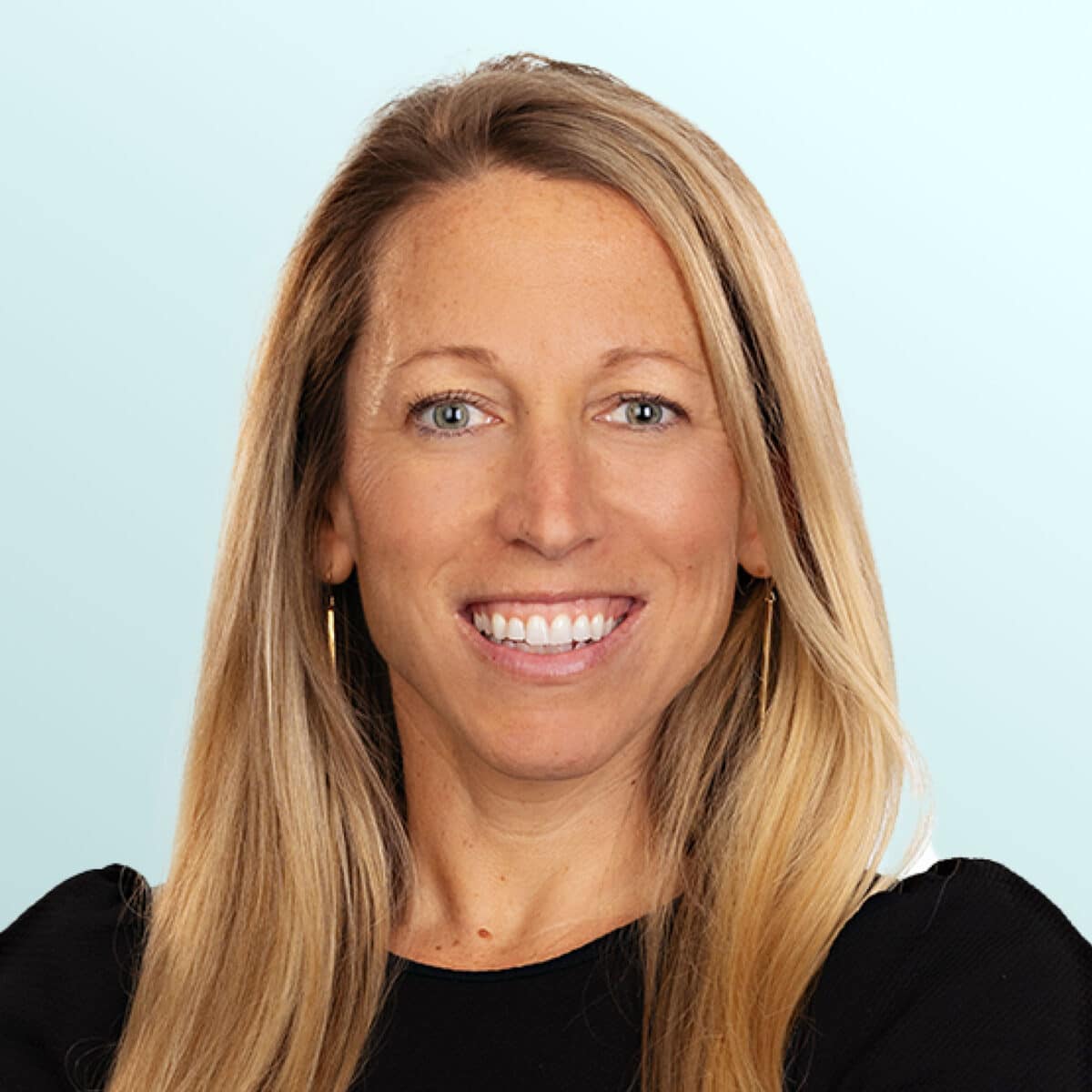 A woman with long blonde hair, wearing a black top and hoop earrings, smiles in front of a light blue background.