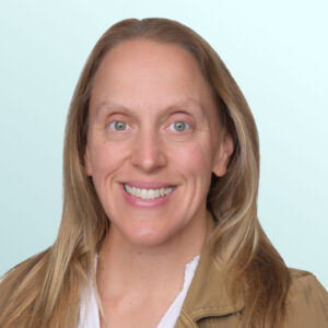 A woman with long light brown hair, wearing a tan jacket over a white blouse, smiles at the camera against a light blue background.