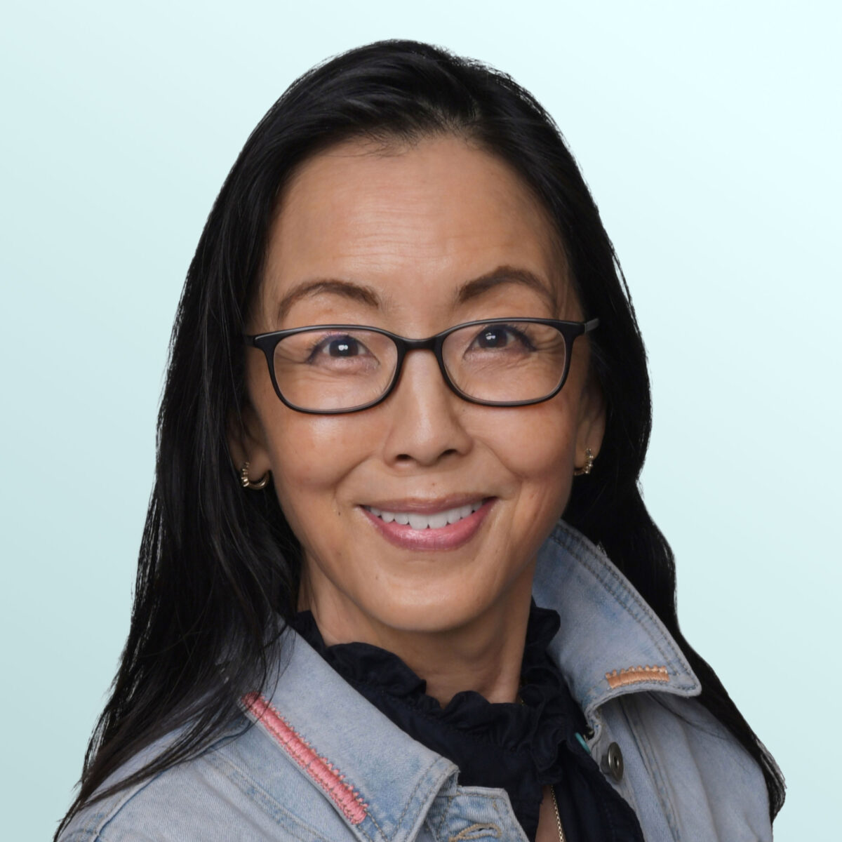 A woman with long dark hair and glasses smiles at the camera. She is wearing a light denim jacket over a black top, standing against a light blue background.
