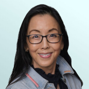 A woman with long dark hair and glasses smiles at the camera. She is wearing a light denim jacket over a black top, standing against a light blue background.