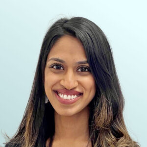 A woman with long, wavy dark hair wearing a black top smiles at the camera against a plain light blue background.