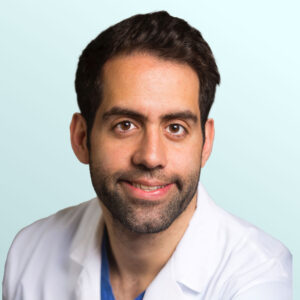A man with short dark hair and a beard, wearing a white medical coat, smiles at the camera against a light blue background.