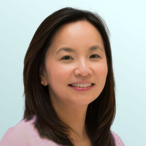 A woman with straight dark hair and a light pink blouse smiles at the camera against a pale blue background.