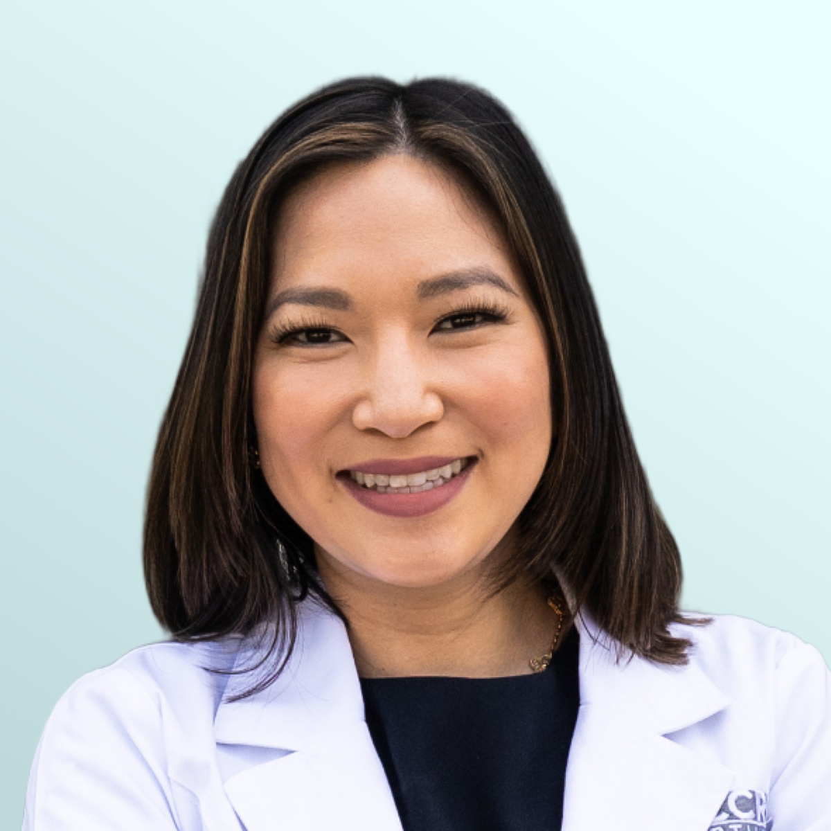 A woman with medium-length dark hair smiles at the camera. She is wearing a maroon top and a delicate necklace with a pendant. The background is a light blue gradient.