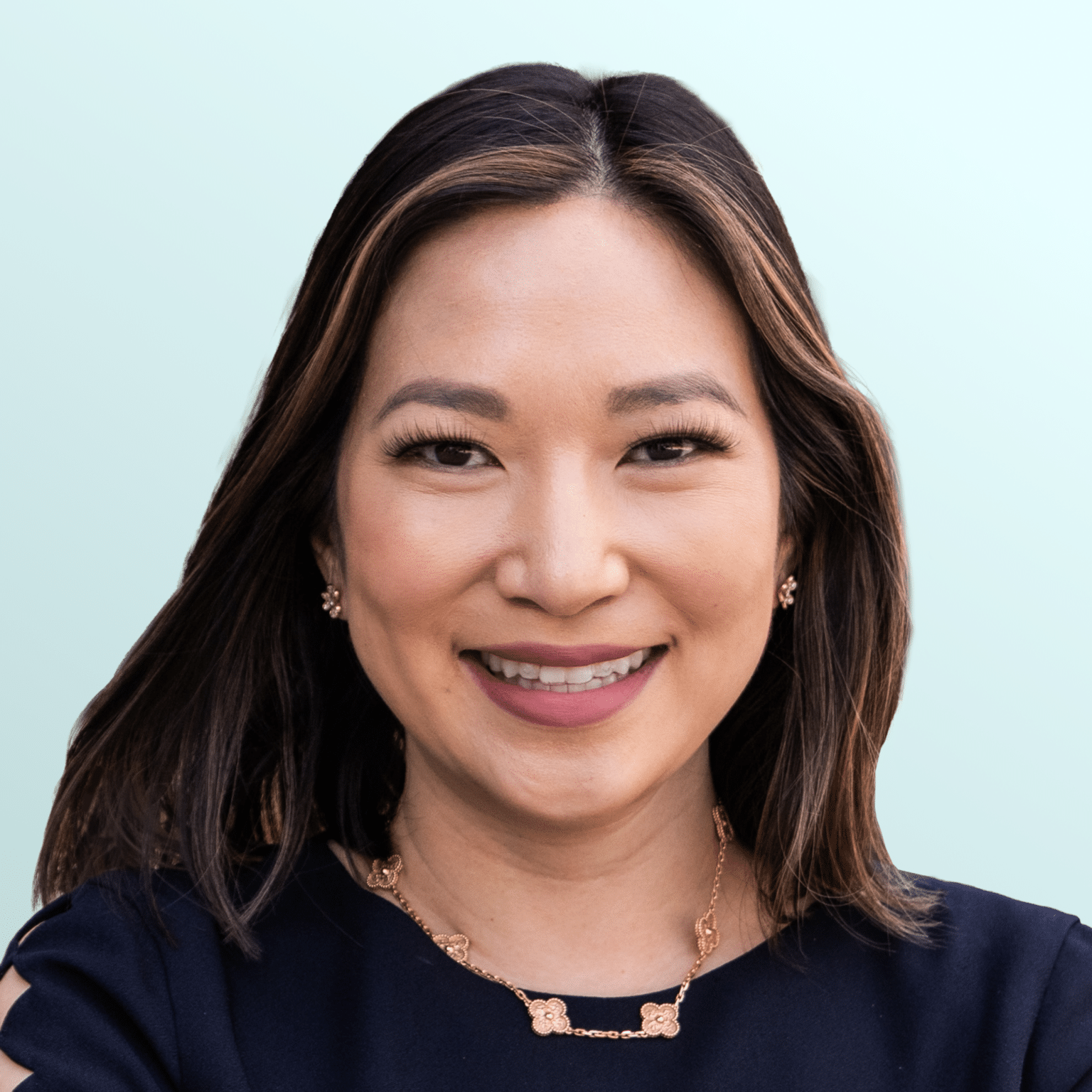 A woman with medium-length dark hair smiles at the camera. She is wearing a maroon top and a delicate necklace with a pendant. The background is a light blue gradient.