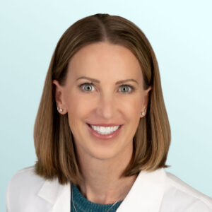 A woman with straight, shoulder-length light brown hair smiles at the camera. She is wearing a white lab coat over a teal top, with stud earrings and layered necklaces. The background is a light blue gradient.