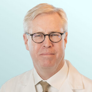 A middle-aged man with blond hair and glasses is smiling. He is wearing a dark suit, white shirt, and a tan patterned tie, with a light blue background behind him.