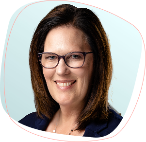A woman with shoulder-length brown hair and glasses smiles at the camera. She is wearing a navy blazer and a delicate necklace, with a light blue background behind her.