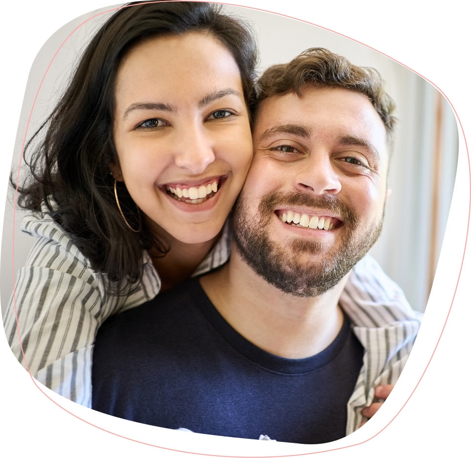 A smiling woman with dark hair hugs a bearded man from behind. Both are wearing casual clothes and looking at the camera, appearing happy and cheerful.
