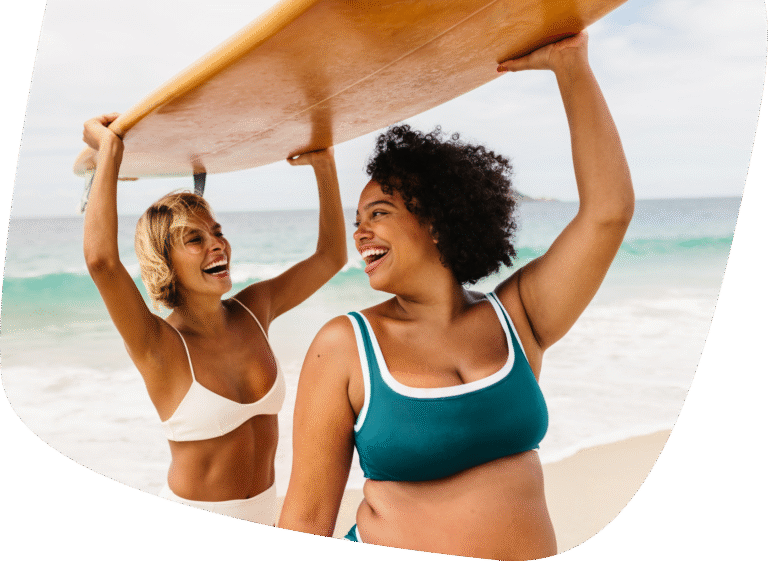 Two women in swimsuits smile and laugh while holding a surfboard above their heads on a sandy beach, with the ocean and sky in the background.