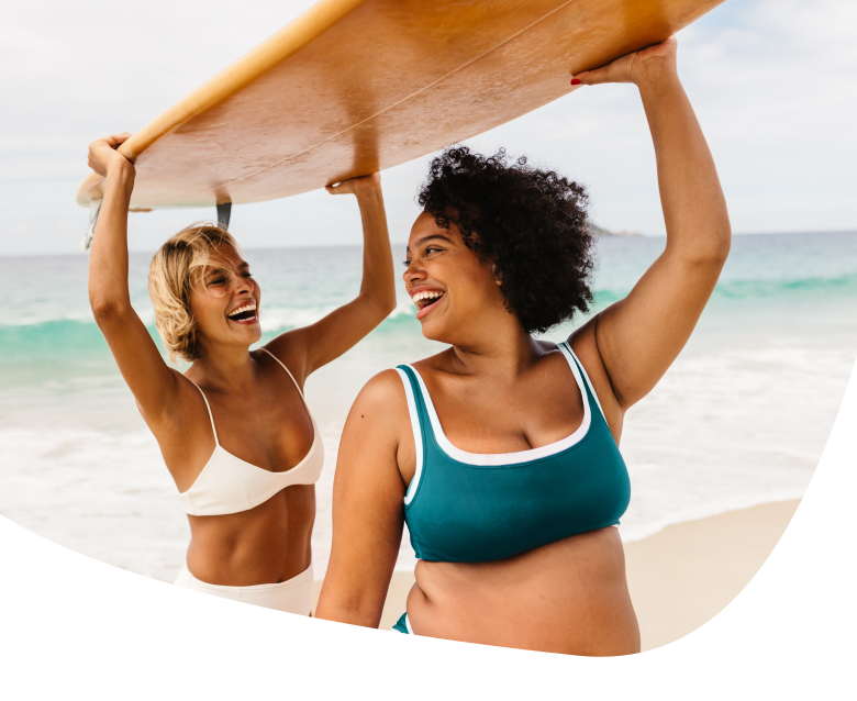 Two women in swimsuits smile and laugh while holding a surfboard above their heads on a sandy beach, with the ocean and waves in the background.