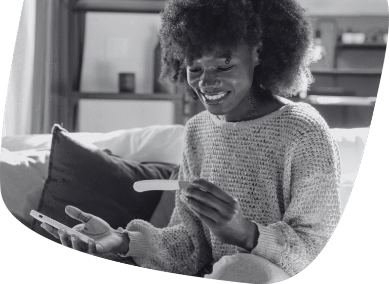 A woman sitting on a couch smiles as she looks at a pregnancy test in one hand and holds a phone in the other, appearing happy and excited.