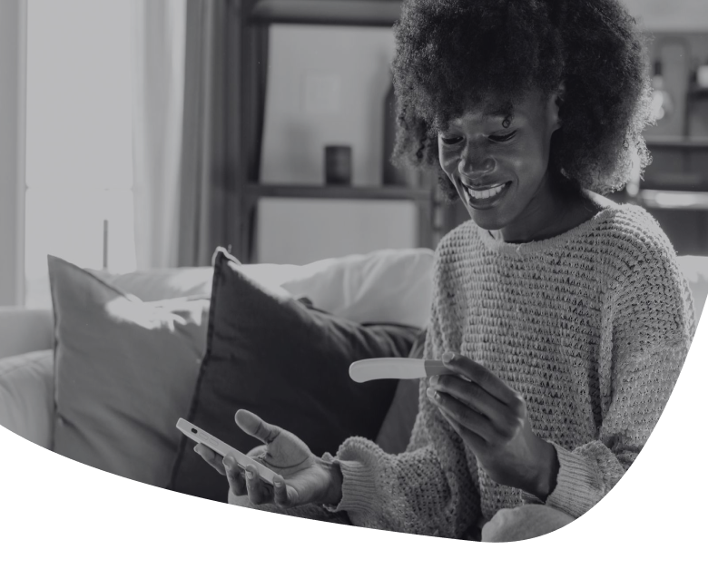 A woman sitting on a couch smiles while holding a pregnancy test in one hand and a phone in the other, appearing happy and hopeful.