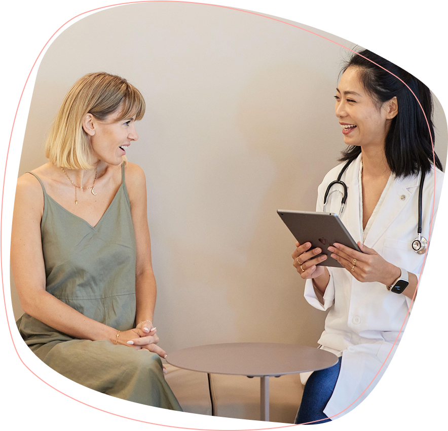 A woman in a green dress sits and talks with a smiling doctor holding a tablet. The doctor wears a white coat and a stethoscope, and they are seated across a small round table in a bright room.