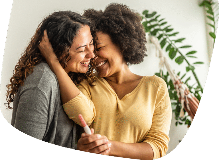 Two women embrace and smile joyfully, with one of them holding a positive pregnancy test. A large green plant is visible in the background, and both appear happy and excited.