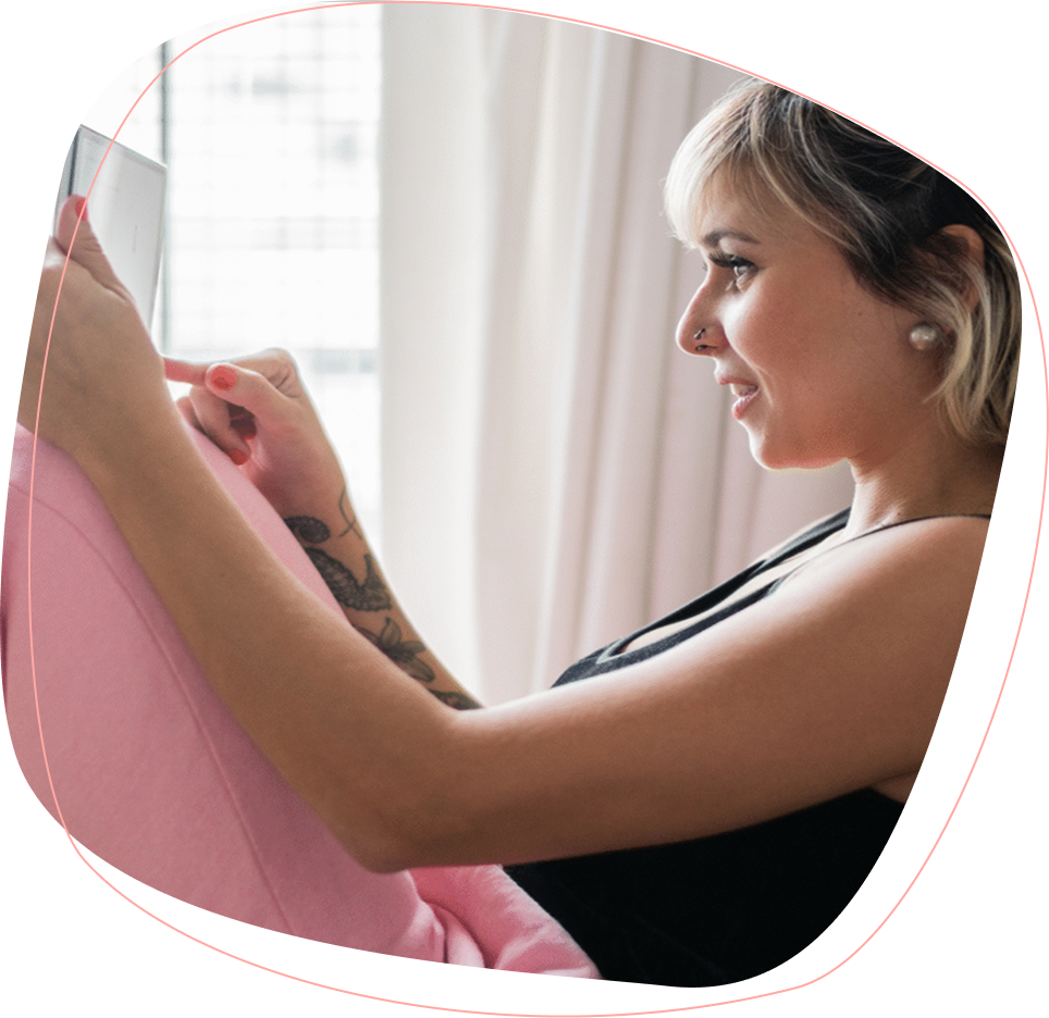 A woman with short blonde hair and a nose piercing sits indoors, smiling while using a tablet. She is wearing a black top and pink pants, and has a tattoo on her forearm. Light filters through curtains in the background.