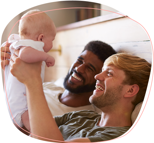 Two men are lying on a bed, smiling and looking up at a baby one of them is holding. The scene is warm and joyful, suggesting a happy family moment.