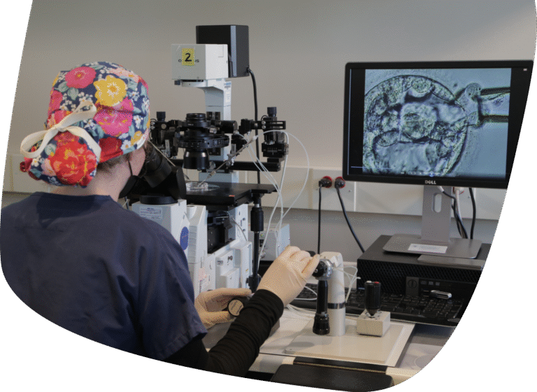 A person in scrubs and a colorful cap uses a microscope and specialized instruments in a laboratory, with a computer displaying a magnified image of cells on the screen.