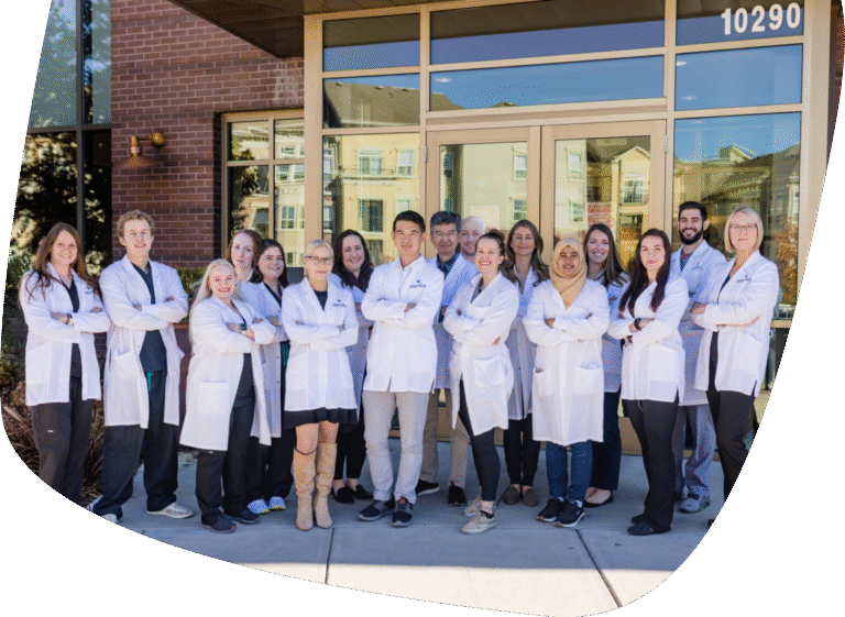 A group of fourteen people wearing white lab coats stand together outside a building with large glass doors and windows, posing for a group photo with arms crossed and smiling.