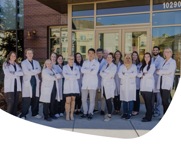 A group of medical professionals wearing white lab coats stand in front of a building entrance, posing together and smiling for a group photo.