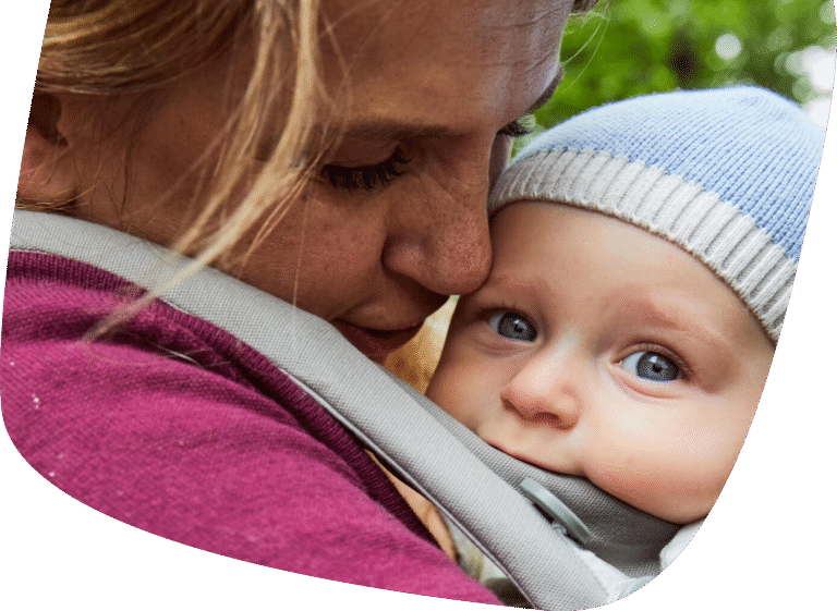 A close-up of a woman lovingly holding a baby in a carrier. The baby, wearing a blue knit hat, looks at the camera with wide blue eyes, while the woman gently presses her face to the babys head.