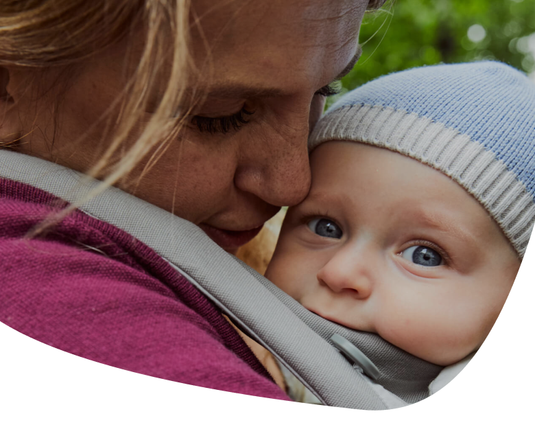 A woman hugs and kisses a baby wearing a blue knit hat and secured in a gray baby carrier. The baby looks at the camera with wide blue eyes. Green foliage is blurred in the background.