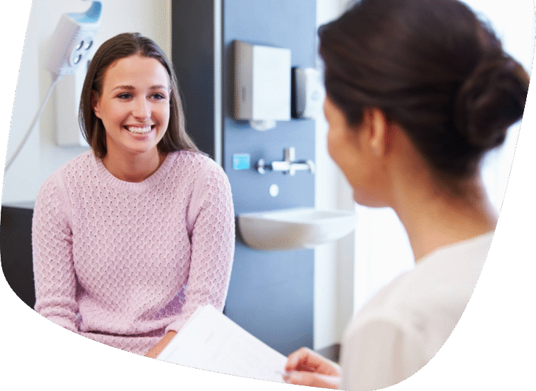A woman in a light pink sweater smiles while sitting across from a healthcare professional, who is holding a clipboard in a medical examination room.