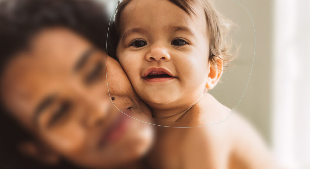 A smiling baby with brown hair hugs an adult, whose face is out of focus. The baby’s face is clearly visible, looking directly at the camera. Soft lighting creates a warm and intimate atmosphere.