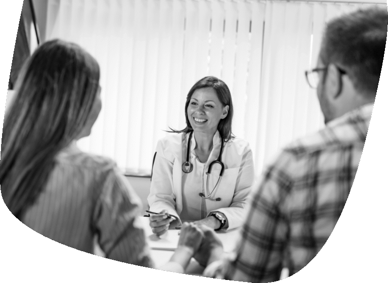 A smiling doctor with a stethoscope around her neck sits at a desk, talking to two patients across from her. The scene is brightly lit with window blinds in the background.
