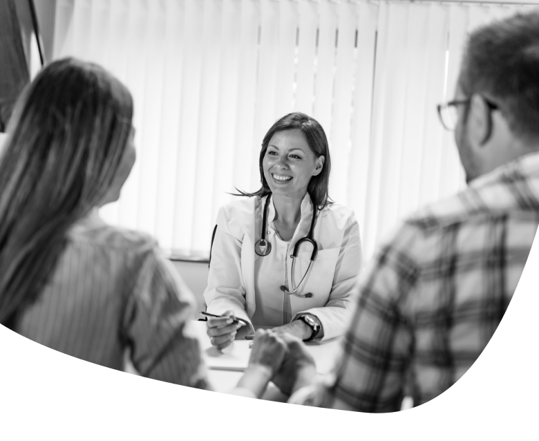 A smiling doctor with a stethoscope sits at a desk, talking to two patients across from her. The scene is in black and white, with light filtering through vertical blinds in the background.