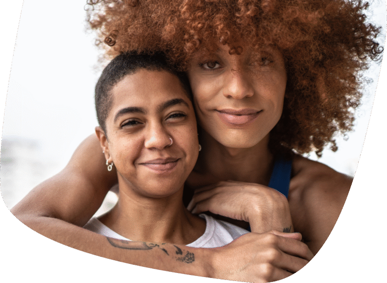 Two people smiling closely together, one with short hair and one with curly, voluminous hair, sharing an affectionate embrace and looking content.