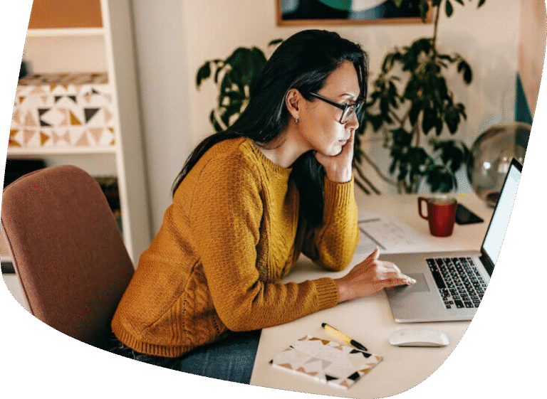A woman wearing glasses and a mustard sweater sits at a desk, focused on her laptop. Papers, a pen, and a red mug are on the desk. There are plants and shelves in the background.