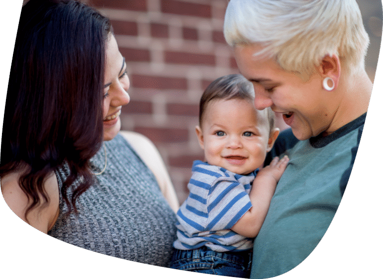 Two adults smile and look at a happy baby in their arms. The baby wears a blue striped shirt, and the three are standing together outdoors in front of a brick wall.
