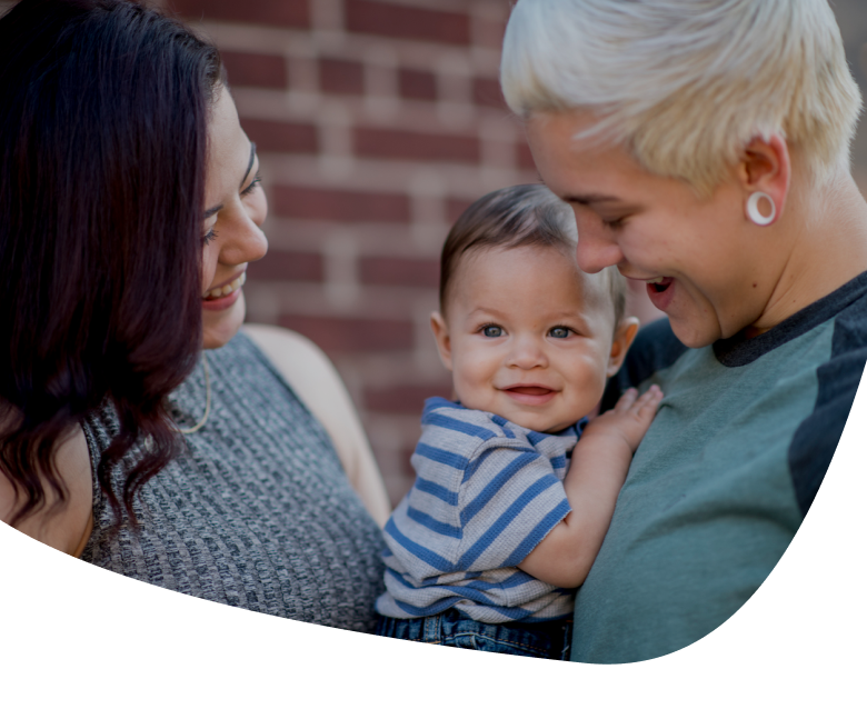 Two adults smiling and holding a happy baby who is looking at the camera, standing outside in front of a brick wall.